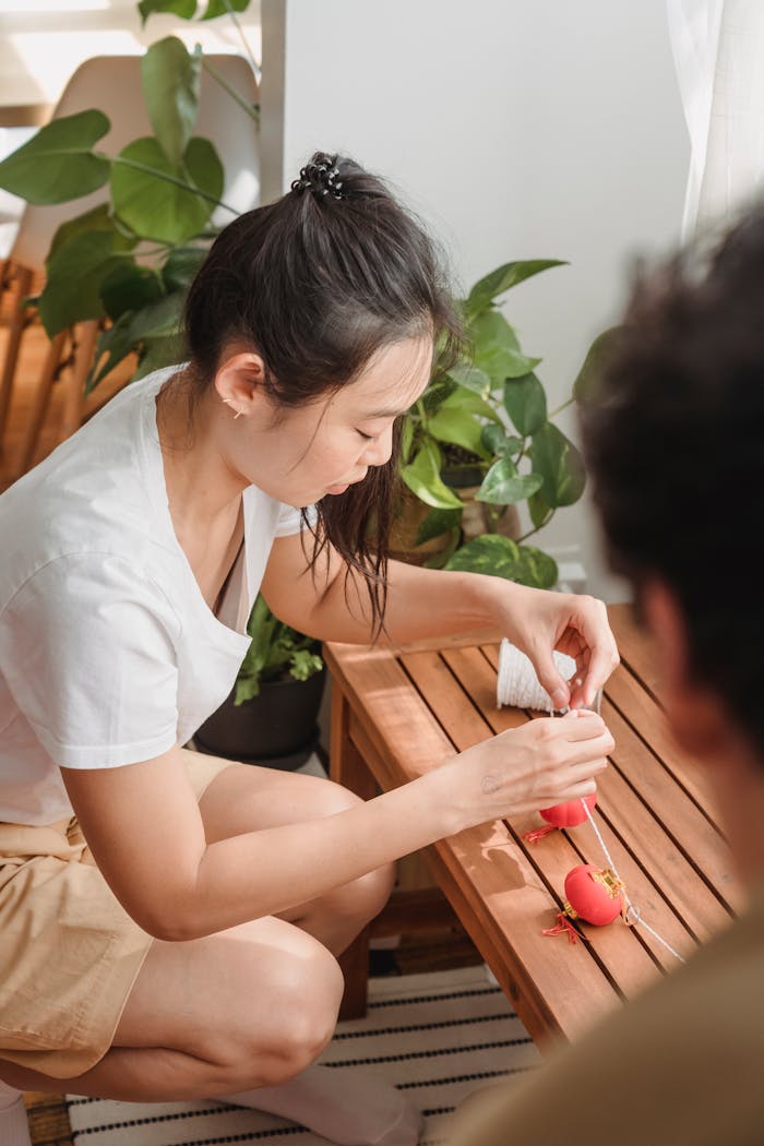 An Asian woman assembles decorative red lanterns at a wooden table indoors, surrounded by plants.